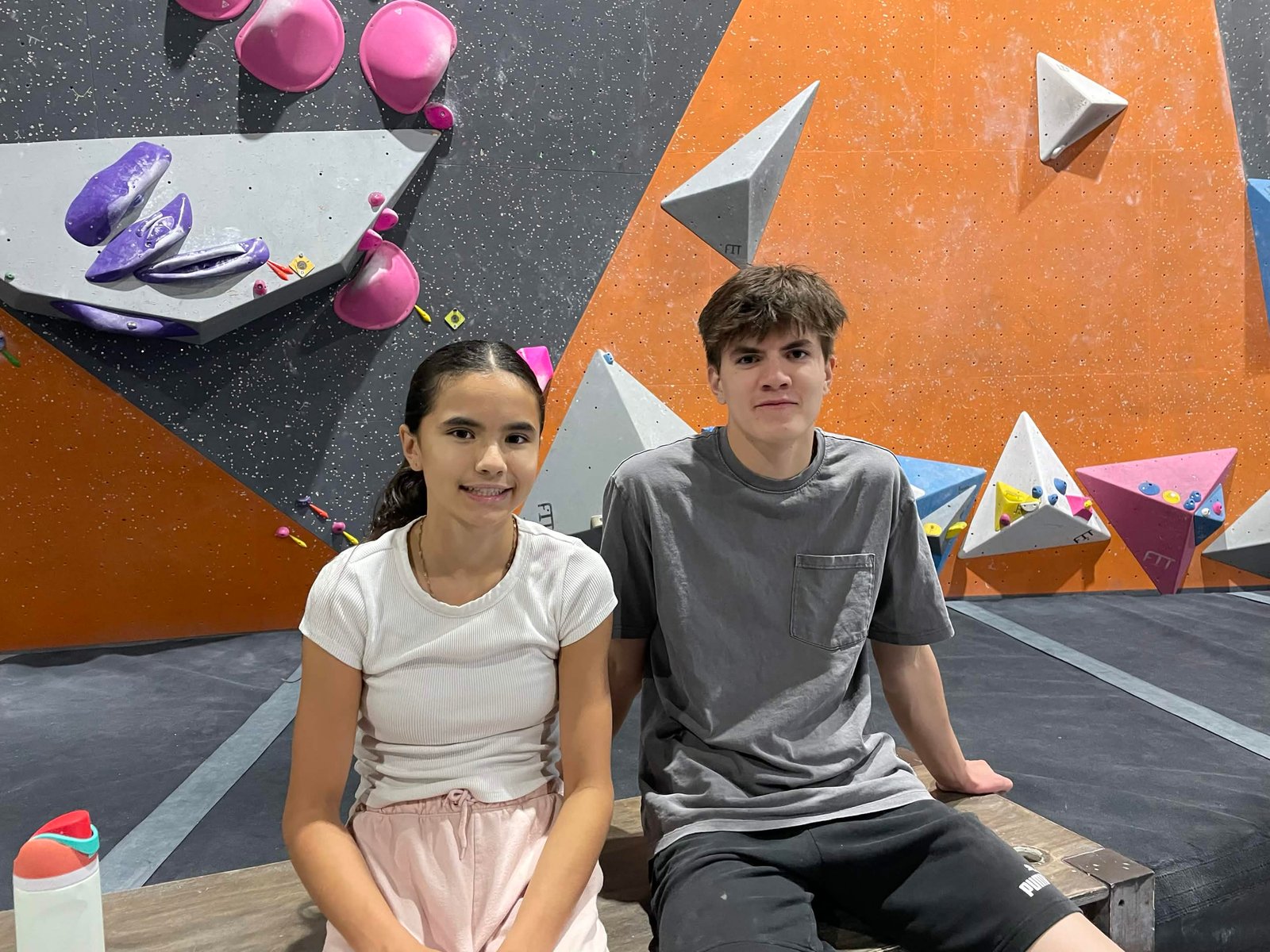 Owen Nitta Mackay sitting on a bouldering mat beside his sister at a climbing gym, colourful holds on the walls behind them.